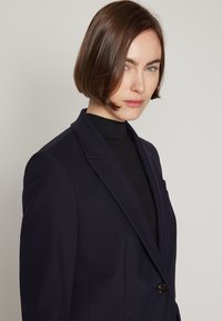 Woman with short brown hair wearing a dark blazer over a black high-neck top, looking slightly to the side against a plain background.