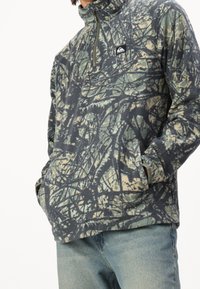 Fleece-pusero tummanvihreänä ja beigenä, jossa on vetoketjukaulus, koko pinnan kattava abstrakti puunoksakuosi ja sivutaskut.