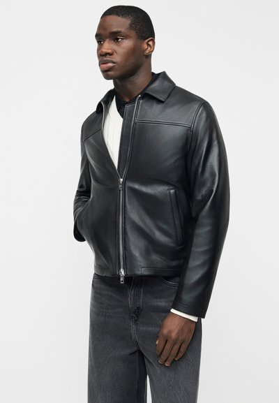 Άνδρας wearing μαύρο δερμάτινο μπουφάν με φερμουάρ, λευκό πουκάμισο και σκούρο τζιν, στέκεται με το ένα χέρι στην τσέπη μπροστά σε απλό φόντο.