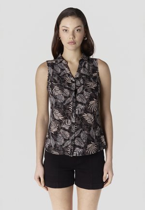 Vrouw draagt een mouwloze zwarte blouse met beige bladpatroon en zwarte korte broek, staand tegen een effen lichte achtergrond.