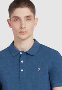 Niebieska koszulka polo uszyta z teksturowanej tkaniny, z trzema guzikami z przodu i pomarańczowym logo po lewej stronie klatki piersiowej.