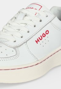 Бяла кожена маратонка с бели дантели, червен "HUGO" лого отстрани, перфорирани детайли и кремава гумена подметка с червен шев.