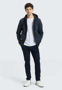 Μπλε ζακέτα με φερμουάρ και διπλό στρώμα κουκούλας, συνδυασμένη με εφαρμοστά σκοτεινά παντελόνια και λευκά αθλητικά παπούτσια. Λείο ύφασμα, άνετο σχέδιο, ελάχιστες λεπτομέρειες.