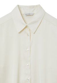 Chemise écrue à boutons avec col pointu et étiquette de marque visible indiquant "CALLIOPE S" à l’intérieur du col.