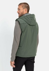 Grüne ärmellose Jacke mit Kapuze, die eine glatte Textur aufweist, und darunter ein graues langärmliges Shirt. Dazu sind schlichte schwarze Hosen sichtbar.