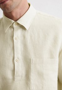 Lichtbeige linnen shirt met een knoopsluiting, puntige kraag en een borstzak. De stof heeft een gestructureerde afwerking.
