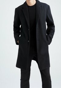 Schwarzer Wollmantel mit langer Silhouette, Reverskragen, zwei Fronttaschen und Ein-Knopf-Verschluss, kombiniert mit schwarzen Hosen.