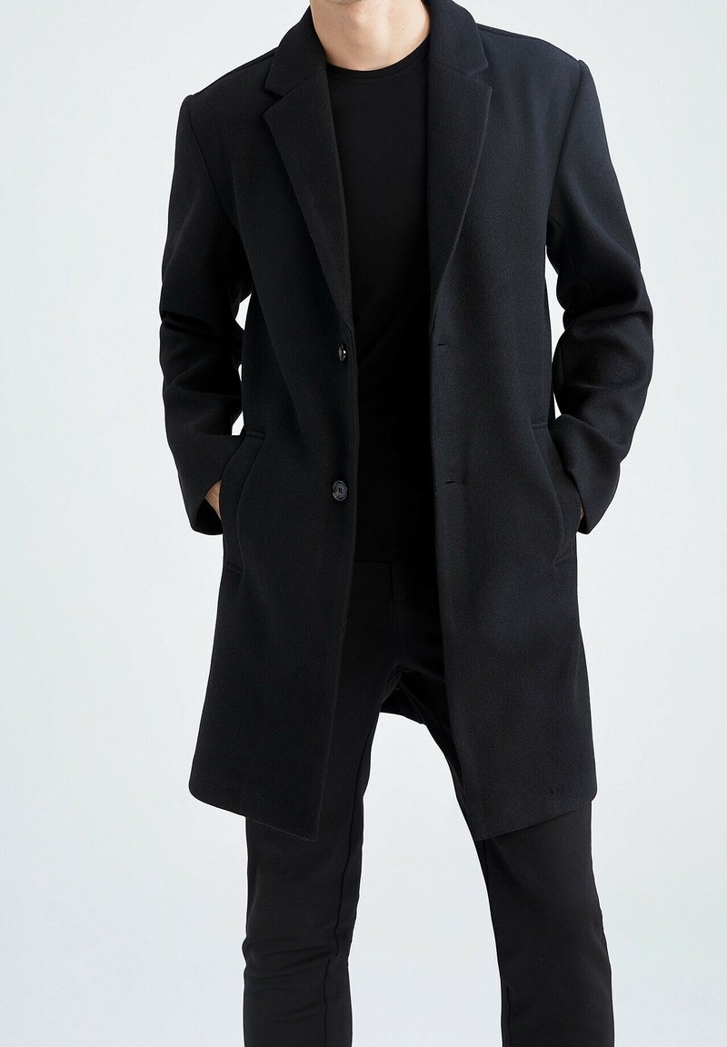 Schwarzer Wollmantel mit langer Silhouette, Reverskragen, zwei Fronttaschen und Ein-Knopf-Verschluss, kombiniert mit schwarzen Hosen.