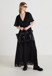 Black, flowing maxi dress with short sleeves, paired with a black leather handbag featuring two front pockets and dual handles. Black shoes complete the look.