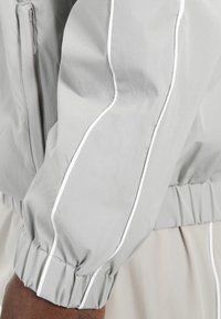 Giacca a vento grigia con polsini elasticizzati, caratterizzata da piping bianco lungo le maniche e una finitura in tessuto testurizzato. Chiusura frontale con zip.