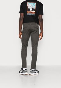 Pantaloni slim-fit verdi scuri con due tasche posteriori, indossati con una maglietta nera con un design grafico. Abbigliamento completato da sneaker nere e grigie.