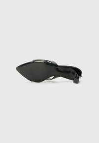 Chaussure à talon haut noire avec un bout pointu, finition brillante, fine bride de cheville et semelle texturée. Présente un petit détail de talon carré.