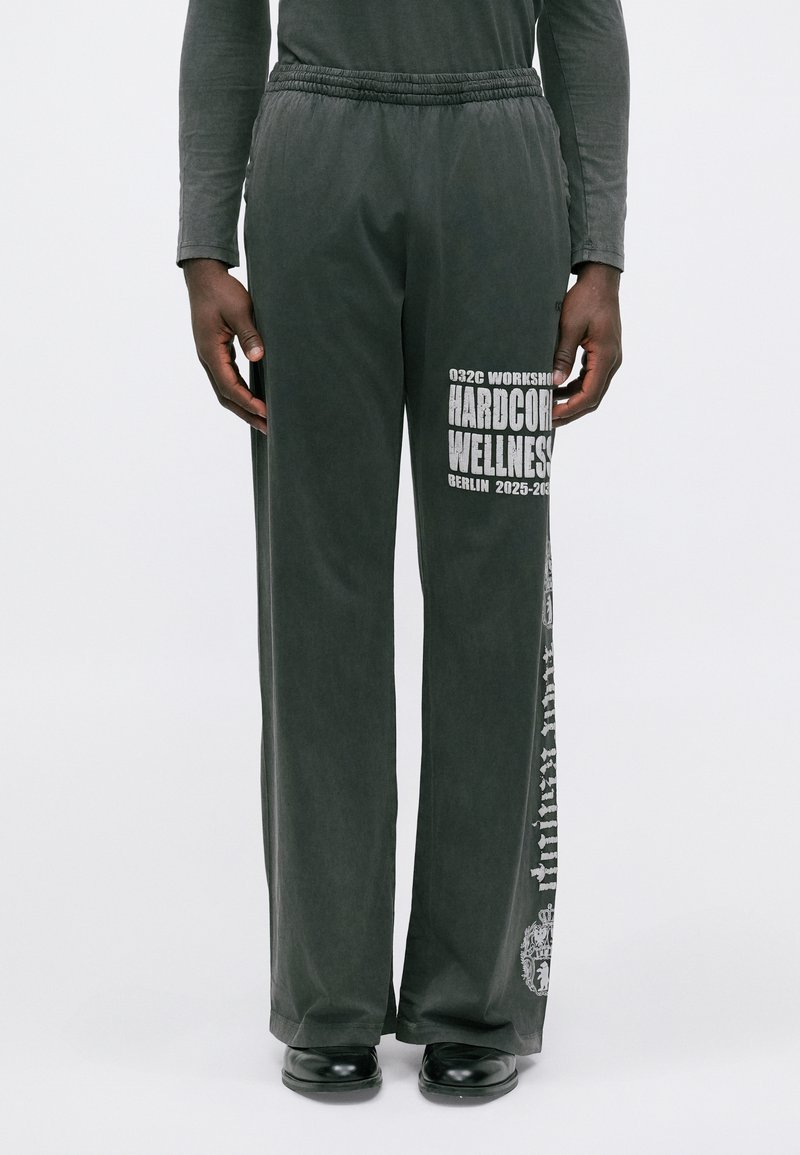 Person i lösa svarta byxor med vit text "HARDCORE WELLNESS Berlin 2025-203x" och svarta skor mot en neutral bakgrund.