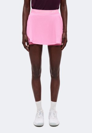Personne portant une jupe de sport rose associée à des chaussettes blanches et des chaussures de sport blanches, debout devant un fond clair uni.