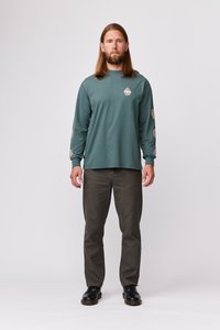 Majica s dolgimi rokavi v teal barvi z osrednjim logotipom in vzorčenimi poudarjenimi rokavi, v kombinaciji s temno sivo hlačami in črnimi čevlji, stoji na belem ozadju.