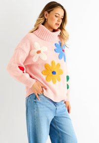Pink knitted sweater with a high collar featuring multicolored floral appliqués in yellow, blue, and white. Denim jeans complete the outfit.