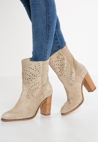 Botas de tobillo de ante beige con patrones recortados en el eje, puntera redonda y tacón de madera apilada. Llevadas con jeans de mezclilla azul.
