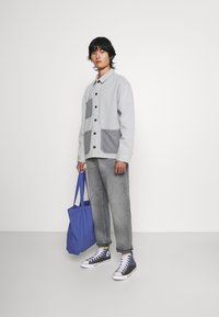 Camisa de denim gris claro con dos bolsillos de parche grises, abotonada, combinada con jeans grises y zapatillas de deporte azul marino. Lleva una bolsa de tote azul.