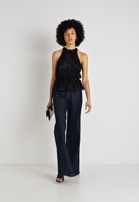 Melnā halter peplum blūze ar ziedu aplikācijām, apvienota ar platām tumšām džinsu biksēm. Modelis tur nelielu melnu klēpjdatoru, stāvot uz neitrāla fona.