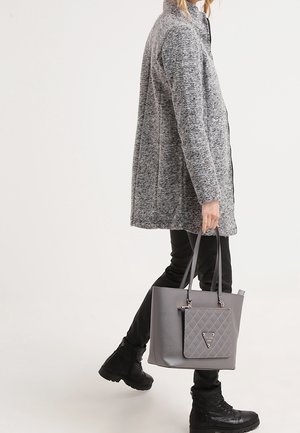 Persona che indossa un cappotto grigio testurizzato e pantaloni neri, che porta una borsa a tracolla grigia imbottita, camminando con un piede leggermente sollevato.