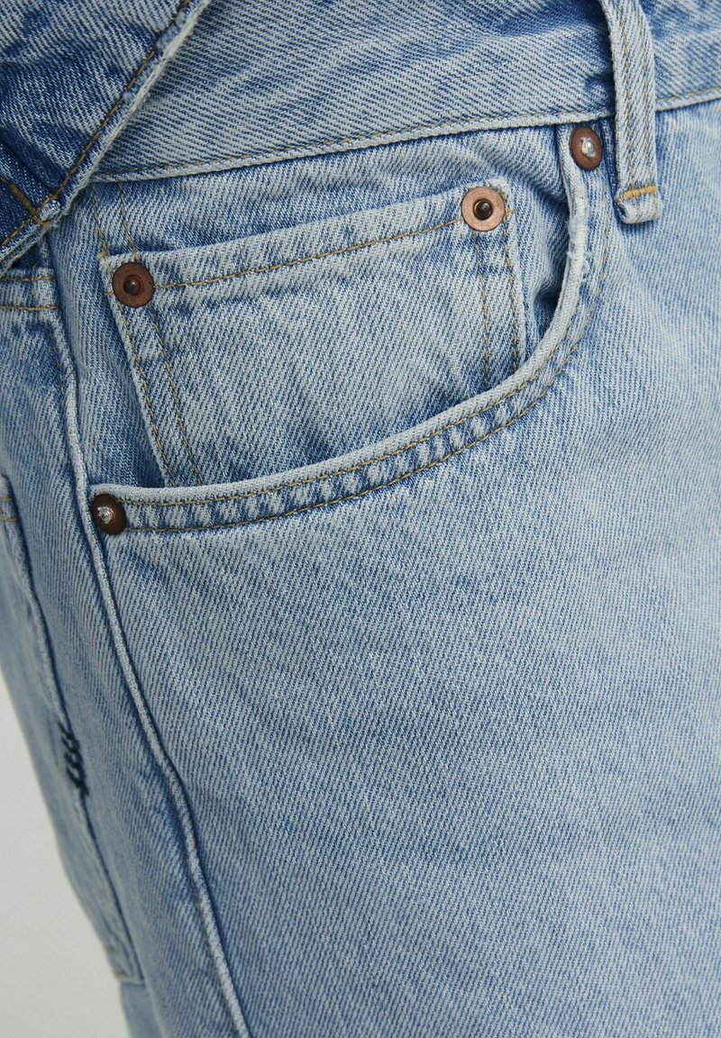 Vaqueros de denim azul claro con un bolsillo frontal adornado con remaches de color cobre y detalles de costura, que presentan una textura desvaída.