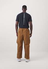 Černé sportovní tričko s nápisem "Under Armour" potisknutým vertikálně na zádech, spárované s hnědými cargo kalhotami. Má elastický pas a boční kapsy.