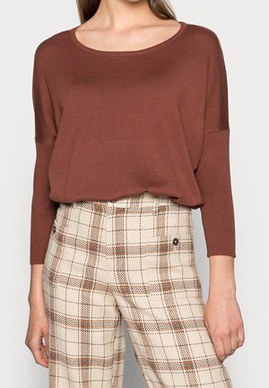 Kvinde iført en løs brun bluse med lange ærmer, stukket ned i beige ternede bukser med høj talje og orange og sorte striber.