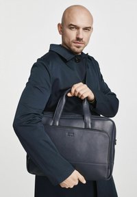 Granatowa skórzana teczka na dokumenty z podwójnymi rączkami, prostokątny kształt, gładka tekstura oraz subtelny detal logo z przodu. Prosty, funkcjonalny design.