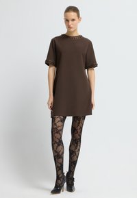 Brą dress z krótkim rękawem w kolorze brązowym z teksturowanym dekoltem i mankietami; zestawiony z czarnymi rajstopami w koronkowy wzór i czarnymi, spiczastymi szpilkami.