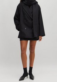 Musta kaksirivinen takki, jossa on korkea kaulus, teksturoitu kangas ja suuret napit, yhdistettynä istuvaan mustaan minimekkoon ja korkeakorkoisiin kenkiin.