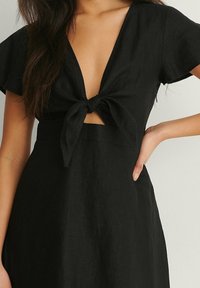Femme portant une robe noire à manches courtes avec un nœud à l'avant, montrant une petite découpe sous la poitrine.