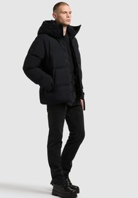 Mustade sulejopedega puffer-jakk, millel on kapuuts, ja millel on tikitud muster ning ees lukuga. Seljas mustad teksad ja mustad kinnised saapad.