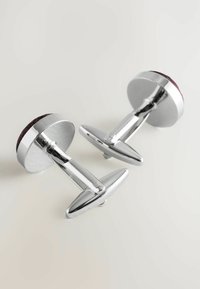 Silberne Manschettenknöpfe mit runden, strukturierten Basen aus tiefrotem Stoff. Elegante, polierte Metallarme führen zu abgerundeten, flachen Enden.