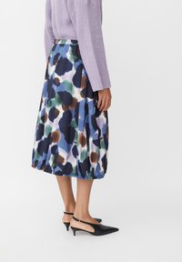 Persona che indossa una gonna midi con motivi astratti blu, verde, marrone e neri, un maglione lavanda e décolleté nere con tacco.