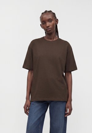 Femme aux cheveux tressés portant un t-shirt marron surdimensionné à manches courtes et un jean bleu, debout sur un fond clair uni.