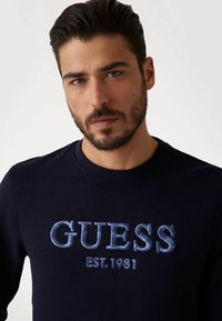 Granatowa bluza z wytłaczanym napisem "GUESS EST. 1981" w jasnoniebieskim kolorze. Wykonana z miękkiego materiału, posiada okrągły dekolt i długie rękawy.