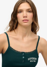 Top de tirantes de canalé verde oscuro con detalle de encaje, que presenta tres botones decorativos y "SUPERDRY NEW YORK" bordado en un color claro.