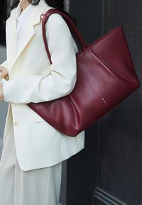 Mujer con traje color marfil sosteniendo un gran bolso tote de cuero burdeos texturizado sobre el hombro.