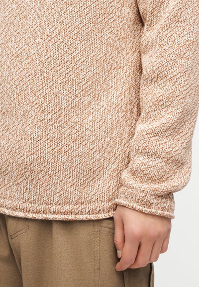 Gros plan d'une personne portant un pull en maille beige et blanc ainsi qu'un pantalon marron, montrant le côté de son torse et une main détendue le long du corps.