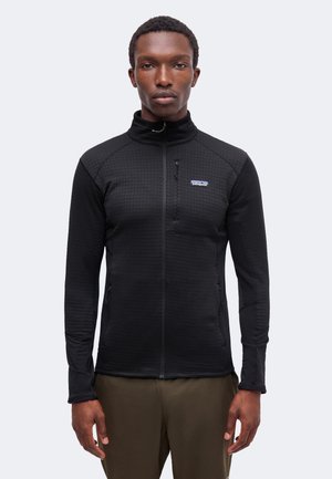 Homme portant une veste noire zippée intégrale avec un petit logo Patagonia, debout devant un fond uni gris clair.