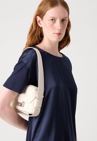 Borsa a spalla off-white con un design elegante e strutturato, dotata di tracolla regolabile, appoggiata su una maglietta blu navy, con un dettaglio di chiusura metallica.