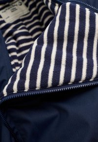 Marineblauwe jas met een gestreepte fleece voering in crème en marineblauw. Expliciete rits met een gladde stoffen buitenkant. Zachte, warme textuur.