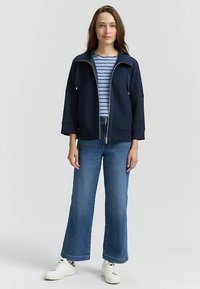 Tamsiai mėlyna užtraukiama striukė su aukšta apykakle, derinama su dryžuotu marškinėliu ir plačiomis mėlynomis džinsais. Aprangą užbaigia balti sportbačiai.