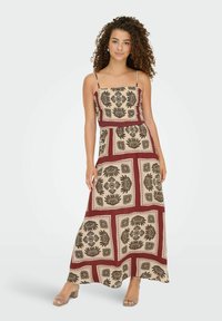 Femme aux cheveux bouclés portant une robe maxi à motifs rouge et beige avec des fines bretelles et des talons ouverts, se tenant contre un fond blanc.