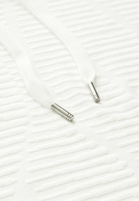 Superficie blanca texturizada con líneas en relieve, presentada con cordones blancos con puntas metálicas plateadas, exhibiendo un diseño limpio y minimalista.