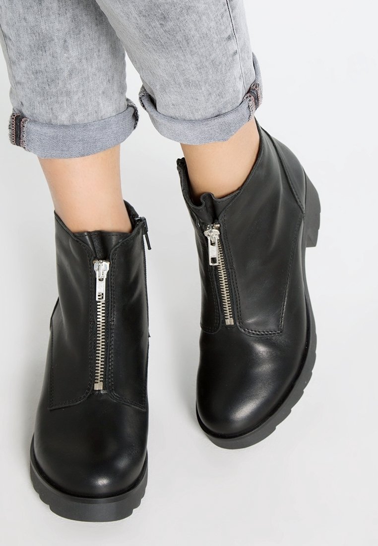 Pieds portant des bottines en cuir noir avec des fermetures éclair argentées à l'avant et un jean gris clair retroussé sur fond blanc.