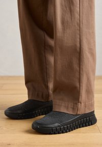 Black slip-on shoes with textured upper and thick sole, paired with loose brown trousers, set on a wooden floor.