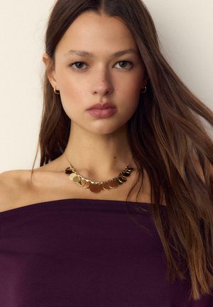 Jeune femme aux longs cheveux bruns portant des boucles d'oreilles créoles en or et un collier pendentif en or, vêtue d'un haut violet foncé décolleté aux épaules.