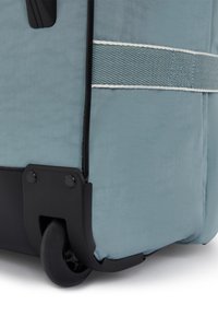 Mjukväska på hjul i dämpad blått tyg med svart plastbas, utrustad med förstärkt hörn, smidig hjul och kontrasterande vit söm.