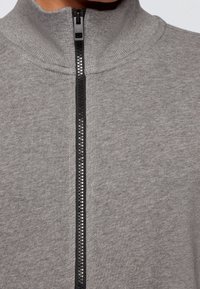 Szary rozpinany sweatshirt z wysokim kołnierzem, z czarnym zamkiem błyskawicznym i teksturowanym materiałem, podkreślającym gładki i miękki wygląd.